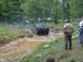 1972 Ford Bronco - A dark-colored off-road vehicle navigates a challenging muddy course with spectators watching from the side.