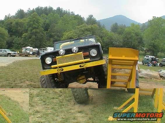 1972 Ford Bronco with yellow bumper and grille guard, angled on uneven terrain. Other vehicles and trees in background