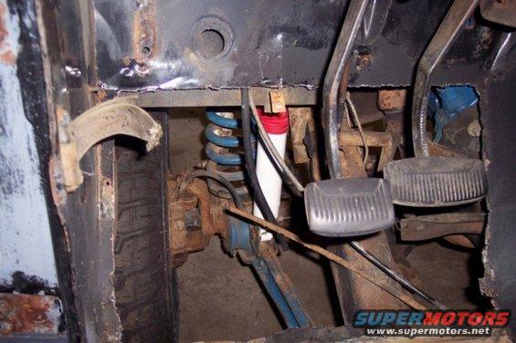 Close-up of rusty interior floor pan, brake and clutch pedals, and front suspension with a blue coil spring and white shock absorber