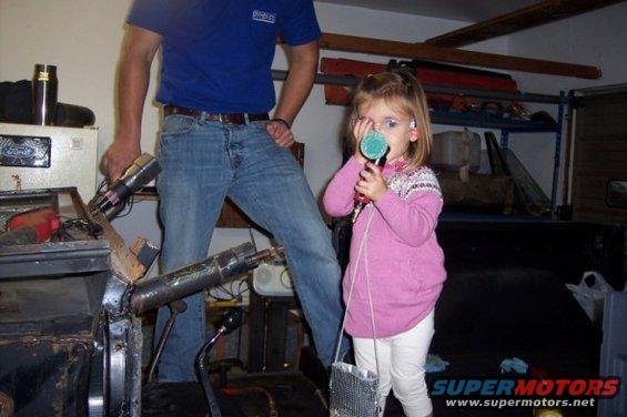Girl holding a microphone in a garage with a man and car parts