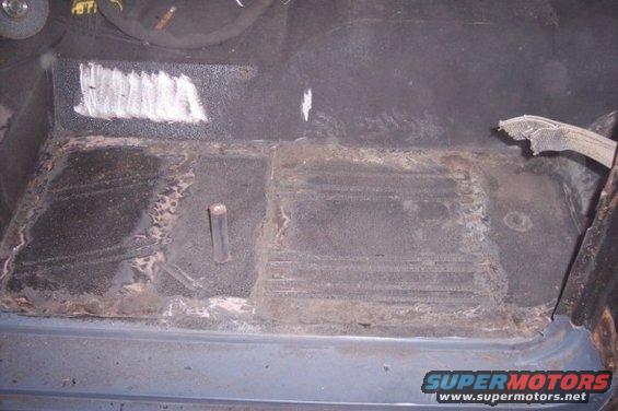 Vehicle floor with rust and white paint marks, showing a metal post and torn fabric