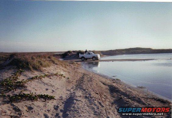 White pickup truck parked near water on a sandy area. A person stands by the driver's side door