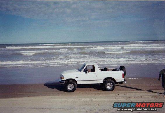 White 1992 Ford Bronco parked on a sandy beach with waves in the background