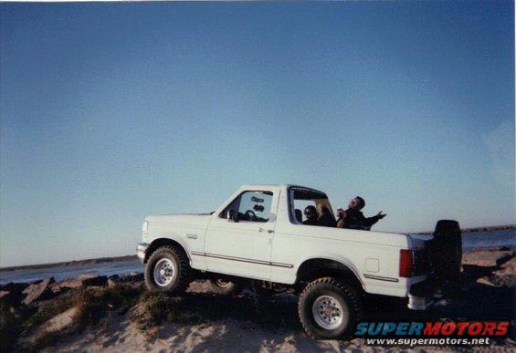 White 1992 Ford Bronco parked on a sandy incline near the ocean with two people in the cab and one in the bed