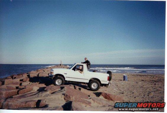 White 1992 Ford Bronco parked on rocks by the ocean, with two people visible