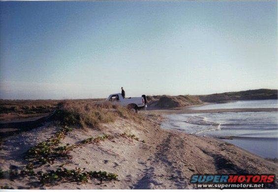 1992 Ford Bronco parked on a sandy dune with a person standing on the roof, ocean in background