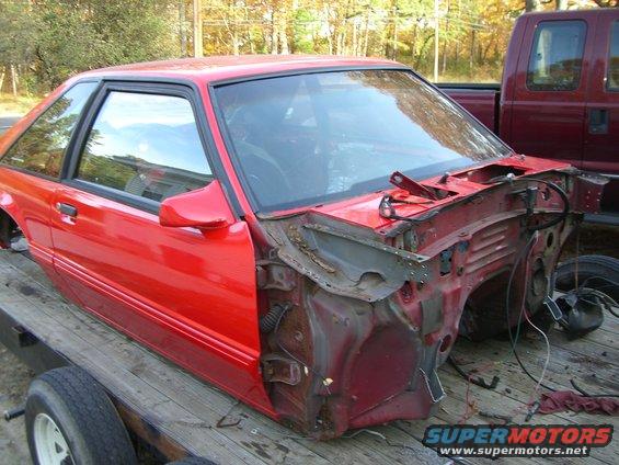 Red car body, likely a Ford Mustang, on a trailer. Front end is dismantled, showing exposed frame and engine bay