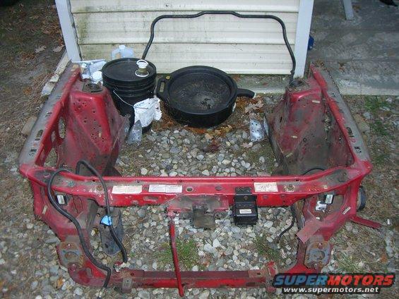 Red car frame with some parts removed, sitting on gravel. Black bucket and oil pan nearby