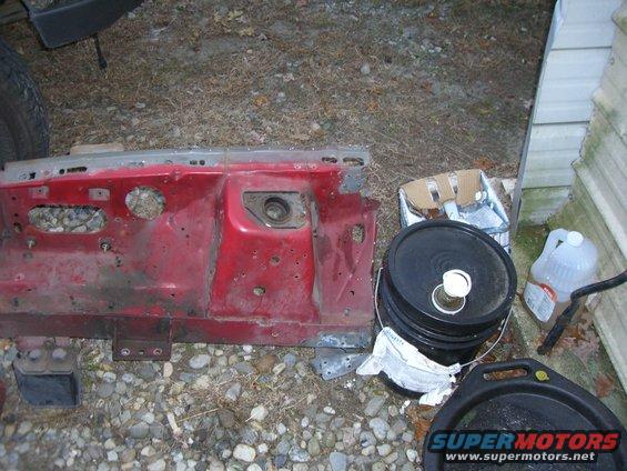 Red metal car part with holes and rust, lying on gravel next to buckets and a jug