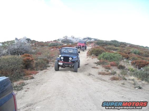 Two Jeeps driving up a dirt road in a hilly, arid landscape