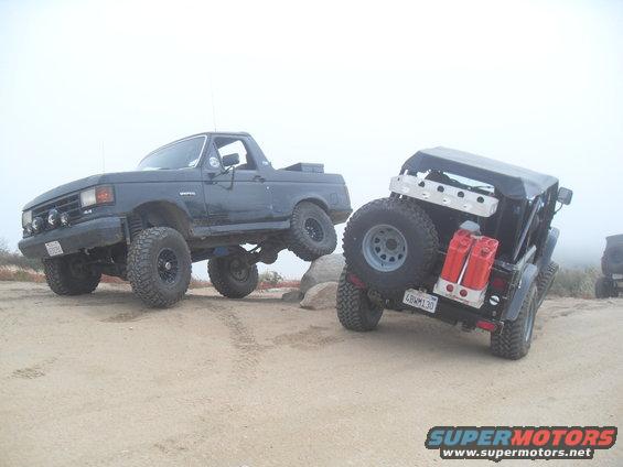 Dark blue SUV with lifted suspension and off-road tires is on rocks. Black Jeep with spare tire and gas cans is next to it
