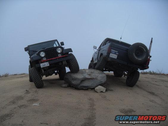 Black 1990 Ford Bronco with spare tire on rear, angled on a rock, with a black Jeep in front