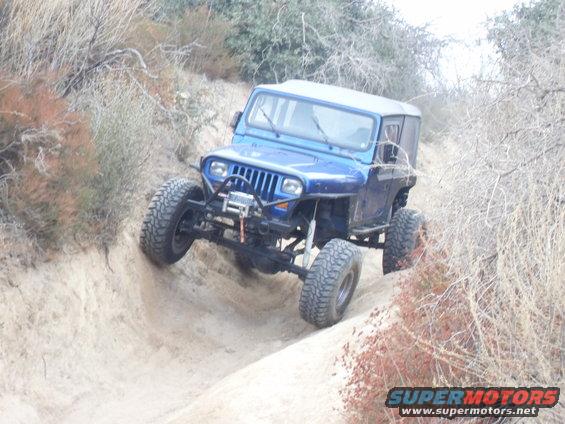 Blue Jeep Wrangler with large tires driving on a dirt path, front winch visible