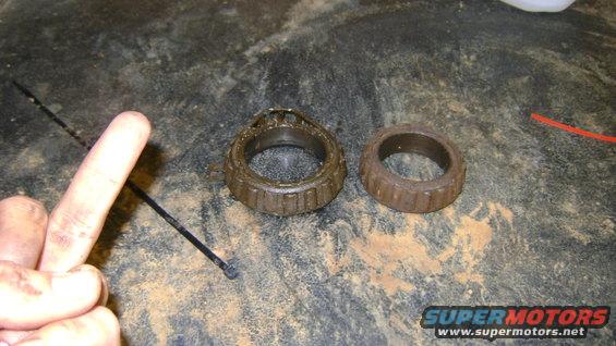 Two used wheel bearings, one with cage damage, on a dirty surface. A finger points to the left