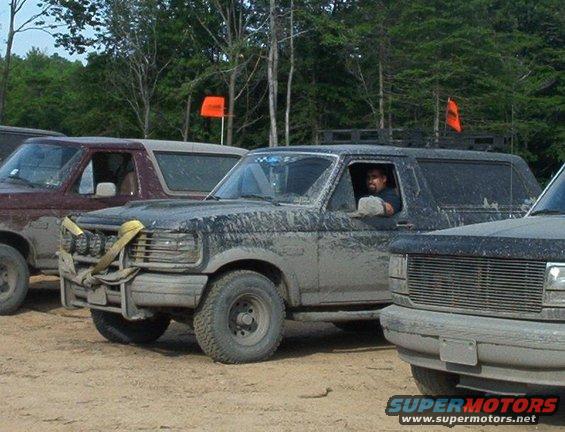 Muddy 1993 Ford Bronco with off-road lights and a driver visible. Another Bronco is to its left, and part of a third vehicle is on the right