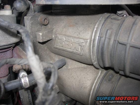 Close-up of a metal engine component with "BBK" embossed on it, connected to a rubber hose