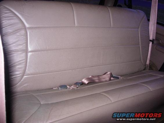 Tan leather rear bench seat with a buckled seatbelt across it. The seat has perforated inserts and visible stitching