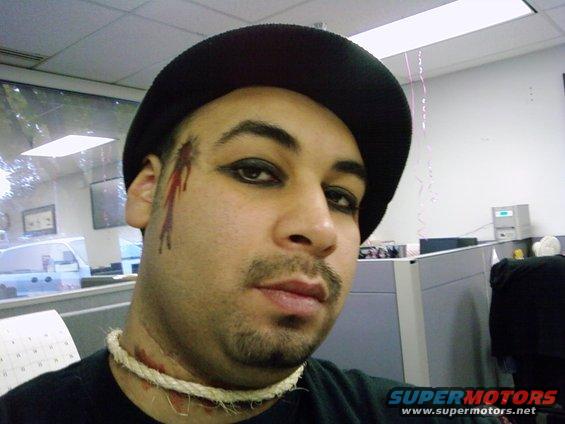 Man with black hat, heavy eye makeup, and fake blood on his face and neck, wearing a rope necklace