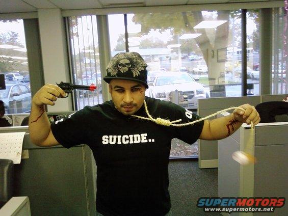Man in office wearing a hat and shirt that says SUICIDE.., holding a toy gun to his head and a noose around his neck