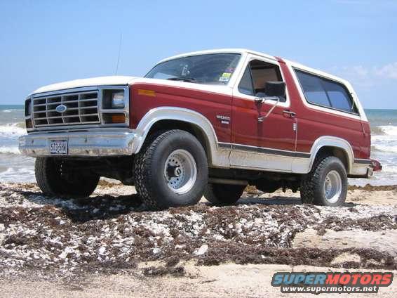 Red and white 1984 Ford Bronco parked on a beach with the ocean in the background