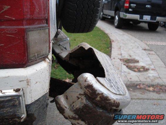 Truck's bent rear bumper and spare tire, red taillight, and white/black body panel. Another black truck is visible in the background