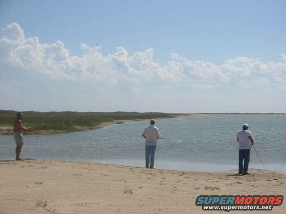 Three men fishing on a beach with a calm body of water and cloudy sky
