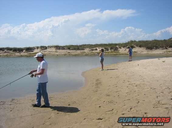 Man in white cap and blue jeans fishes on beach; two other people fish in distance