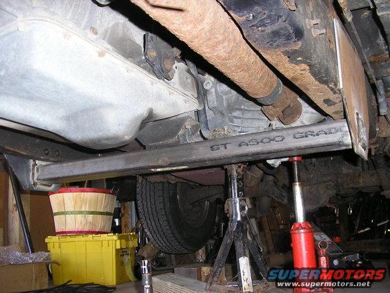 Vehicle underbody with jack stands and a hydraulic jack supporting a metal beam. A tire is visible