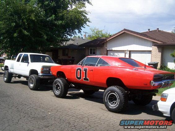 Red 1969 Dodge Charger with "01" on doors, lifted on large tires, parked next to a white pickup truck