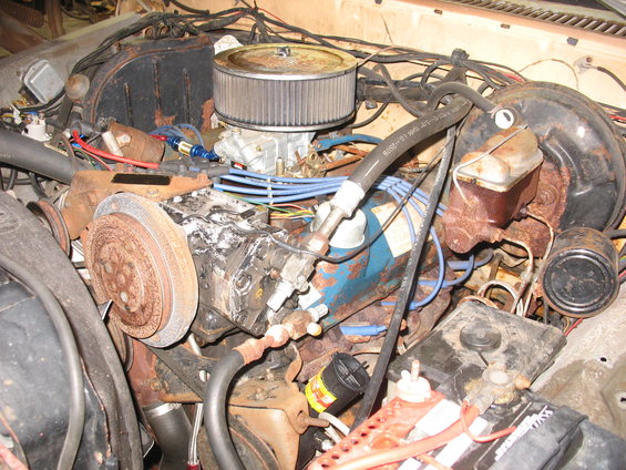 Engine bay with a V8 engine, air filter, carburetor, and various hoses and wires. The engine appears old and rusty