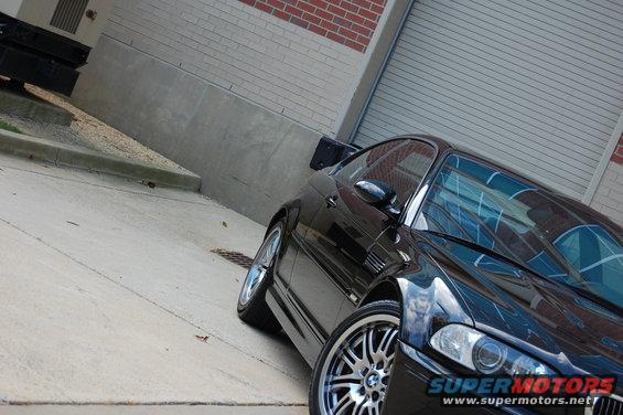 Black BMW M3 parked on concrete, brick wall and garage door in background. Front wheel and side profile visible