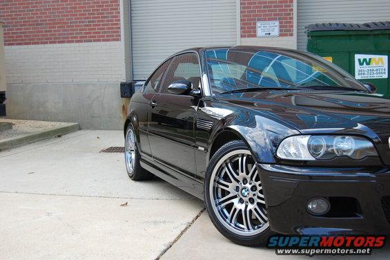 Black BMW M3 coupe, front 3/4 view, parked on concrete near brick building and dumpsters