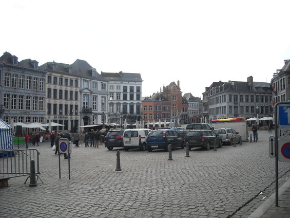 European town square with cobblestone pavement, parked cars, and historic buildings under an overcast sky