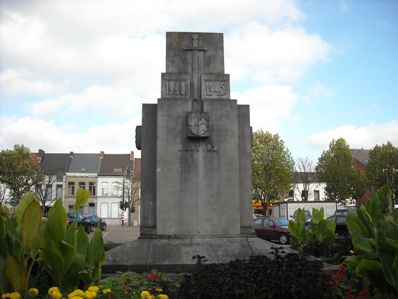 War memorial with dates 1940 and 1945, cars parked in background, residential buildings, and plants in foreground