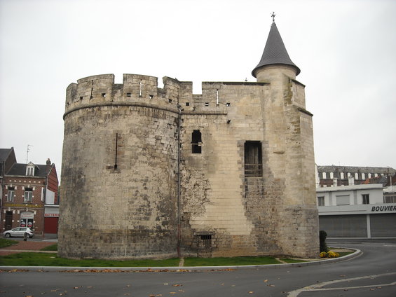 Stone tower with crenellations and a conical roof, next to modern buildings and a street