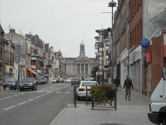 Street in a European city with a large building in the background, cars on the road, and a woman walking on the sidewalk