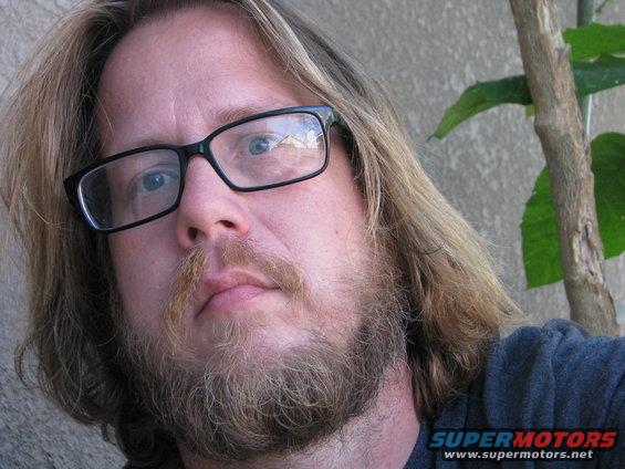 Man with glasses and beard, with a plant and wall in the background. Supermotors logo visible