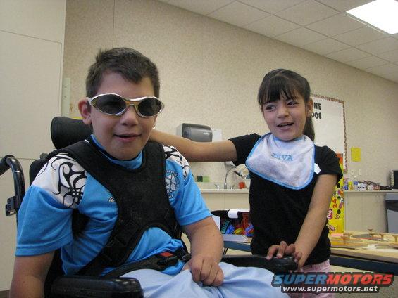 Boy in wheelchair wearing sunglasses and a blue shirt with a black vest, next to a girl wearing a bib
