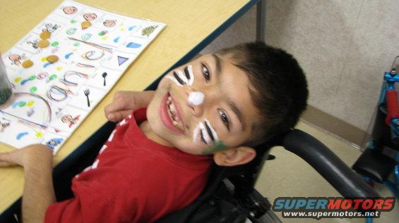 Child with face paint, wearing a red shirt, sitting in a wheelchair next to a table with a chart