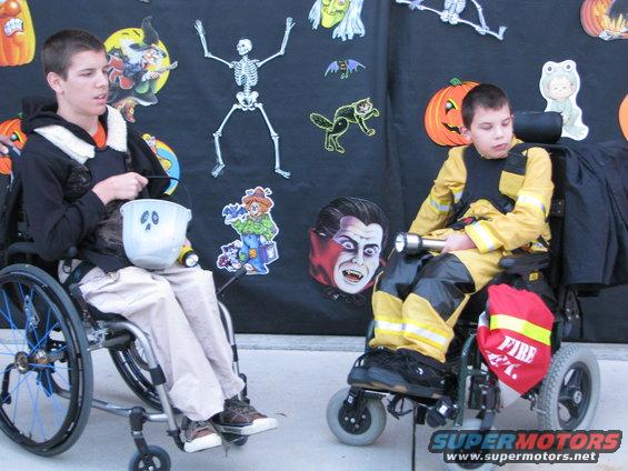 two boys in wheelchairs, one in a black hoodie holding a bucket, the other in a firefighter costume, in front of Halloween decorations