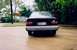 1989 Ford Escort - The rear of a black 1989 Ford Escort sedan is visible on a wet paved surface with trees and foliage in the background.
