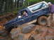 1989 Ford Bronco - A Ford Bronco maneuvers over large rocks and mud on an off-road trail, with two people inside and beside the vehicle.