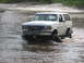 1989 Ford Bronco - A white Ford Bronco navigates through a shallow body of water, kicking up mud.
