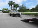 1989 Ford Bronco - A person pushes a lifted white Ford Bronco towards a trailer in a parking lot with palm trees.