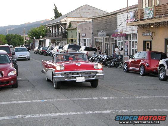 Red convertible driving down a street with a red PT Cruiser and other cars parked