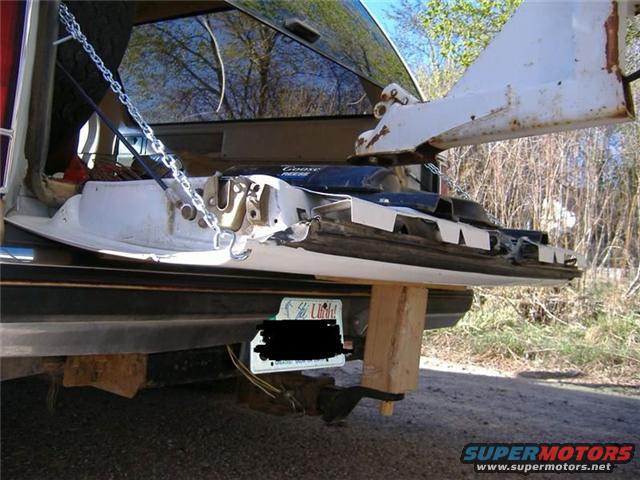 Rear of a vehicle with a tow hitch, chains, and trailer wiring. A wooden block supports the bumper