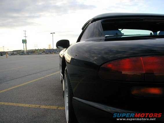 Black sports car parked in a lot, showing rear quarter panel and taillight. The website SUPERMOTORS.NET is visible