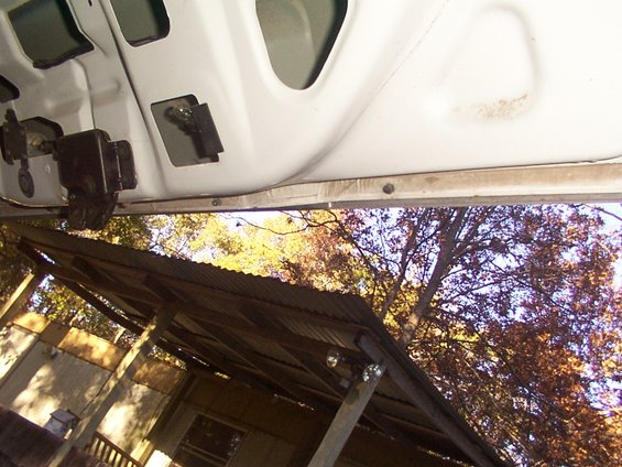 Close-up of a car's open door frame showing the latch mechanism and exterior view of trees and a shed