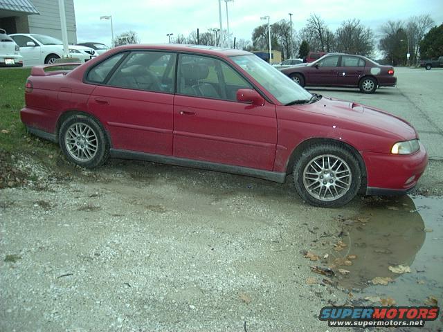 Red sedan parked on gravel with puddles, side view showing front fender and door