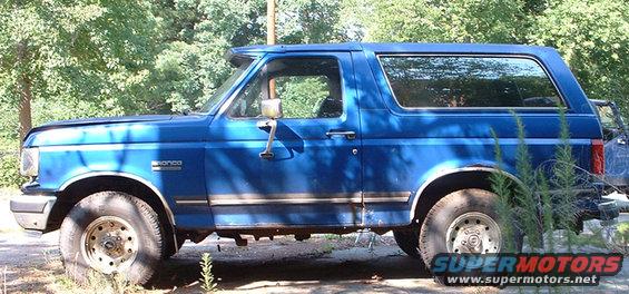 Blue Ford Bronco parked outside with trees in the background. Has a sun visor above the windshield